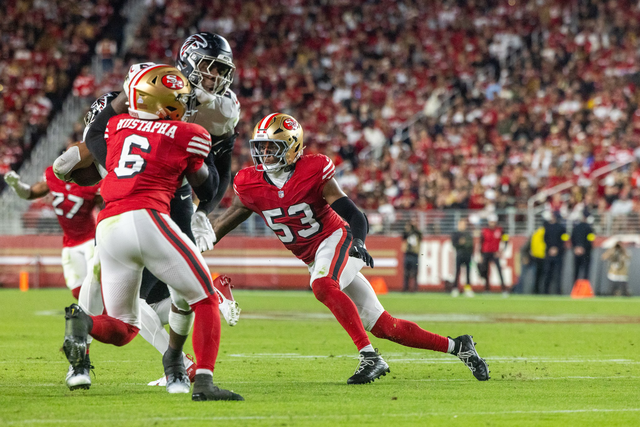 Two San Francisco 49ers defenders wearing red and gold uniforms pursue a Falcons player in black and white, with a crowded stadium in the background.